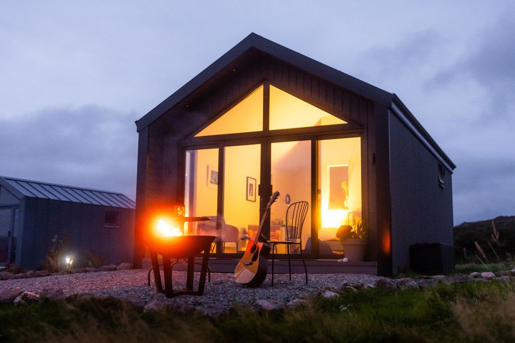 Cabin windows glowing at dusk with a fire burning outside